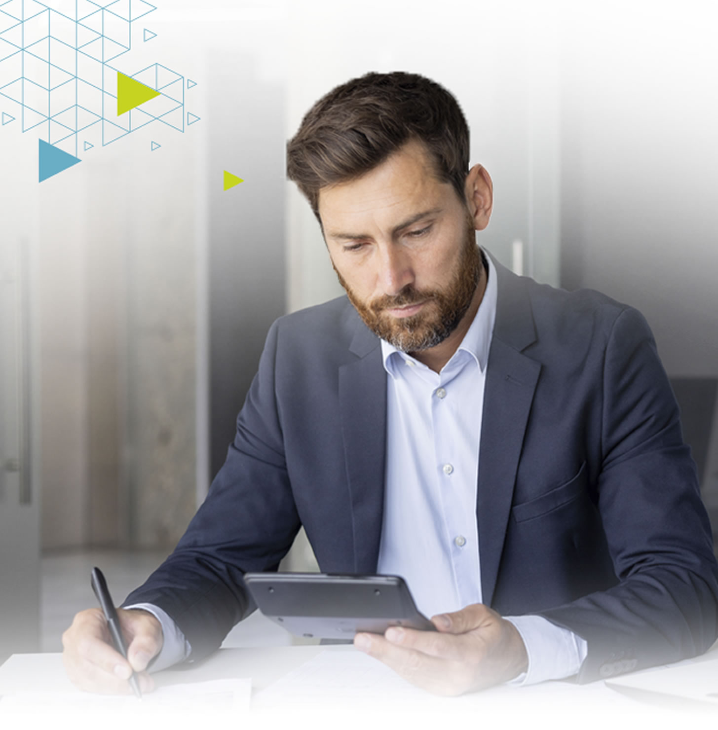 A man in a suit is sitting at a table, using a tablet and writing with a pen. He appears focused and engaged.