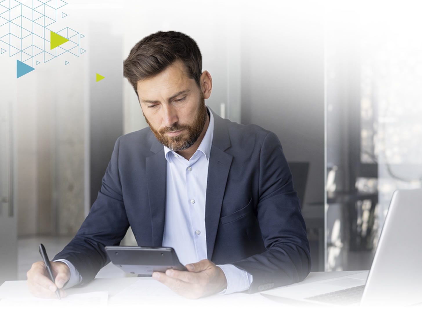A man in a suit sits at a desk, using a tablet and writing with a pen, focused on his work in a modern office setting.