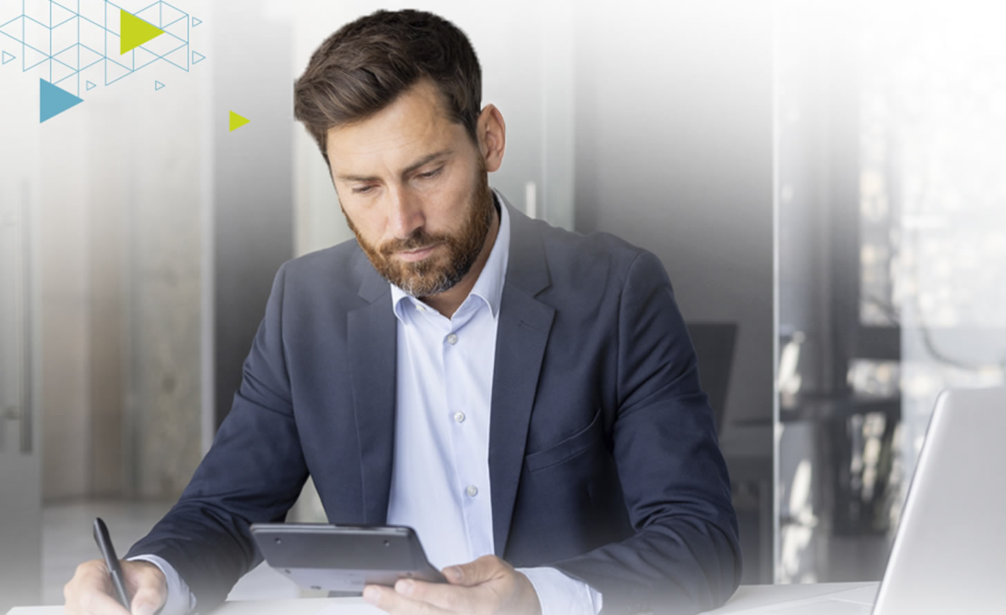 A man in a suit is focused on a tablet while sitting at a desk, with a laptop nearby and a modern office background.